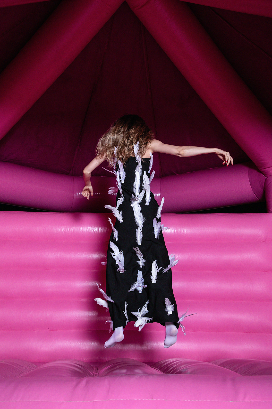 Model in black dress adorned with white feathers posing mid-air in a bright pink bounce house